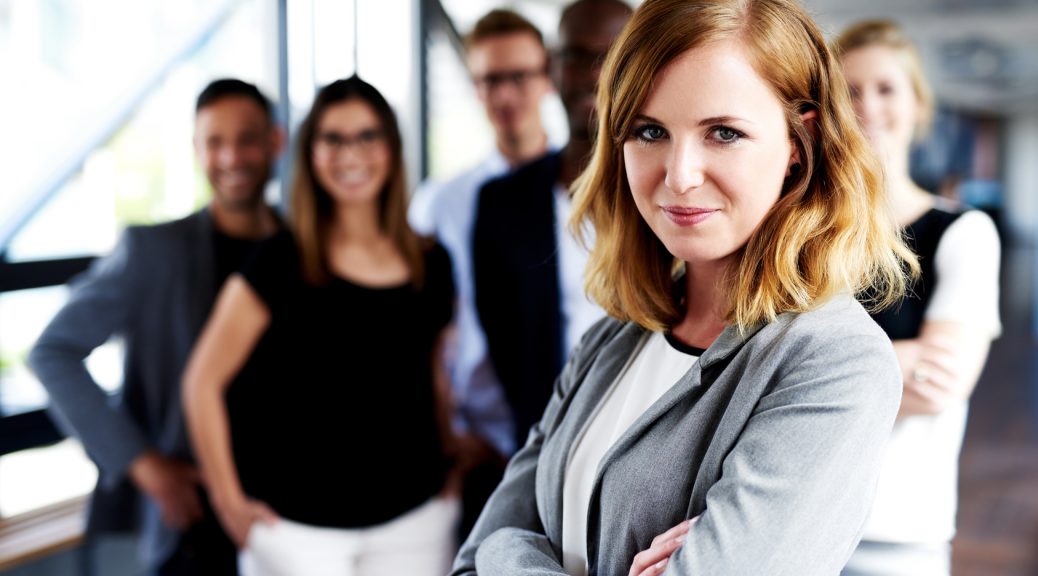 Young white female executive with arms crossed standing in front of colleagues grinning at camera