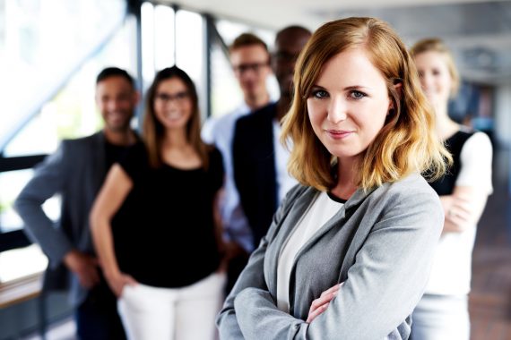 Young white female executive with arms crossed standing in front of colleagues grinning at camera